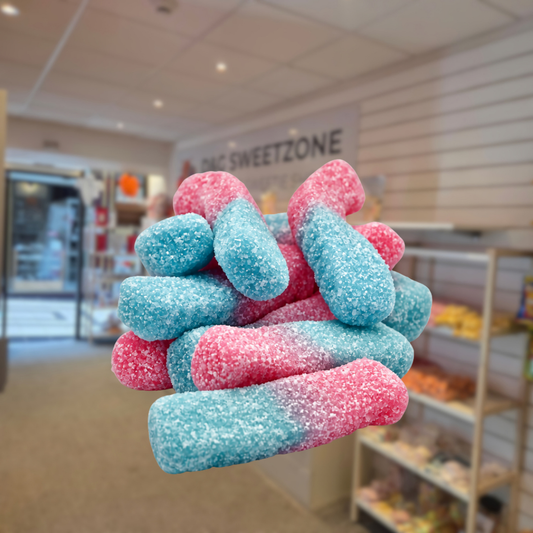 A pile of sugar-dusted gummy candies in pink and blue, shown in front of a blurred bakery shop background with a 'C Sweetzone' sign visible.