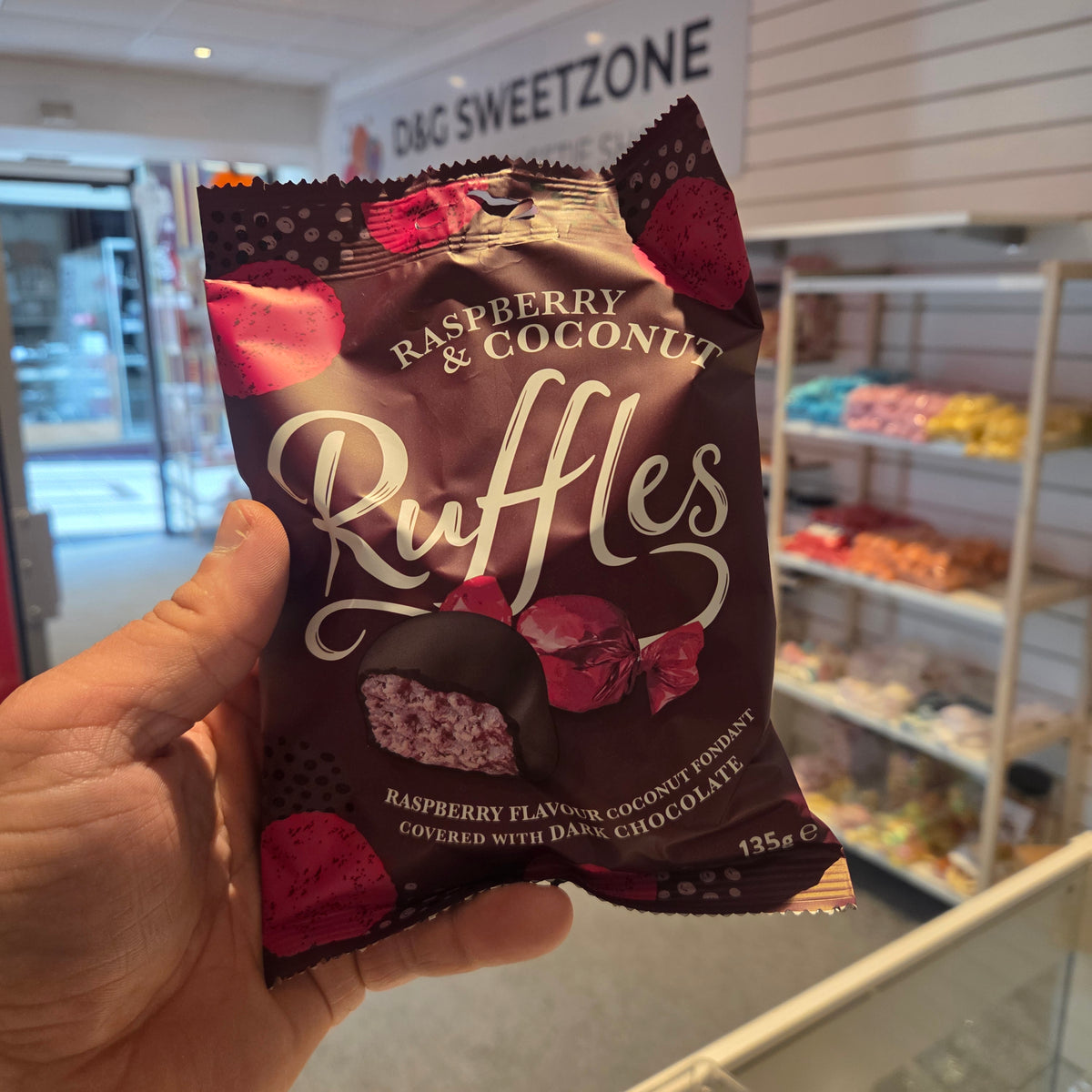A hand holding a bag of Raspberry & Coconut Ruffles chocolate in a shop setting with a D&G Sweetzone sign in the background.